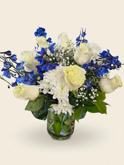 White roses, blue delphinium, white chrysanthemums and baby's breath arranged in a clear glass vase on a cream background