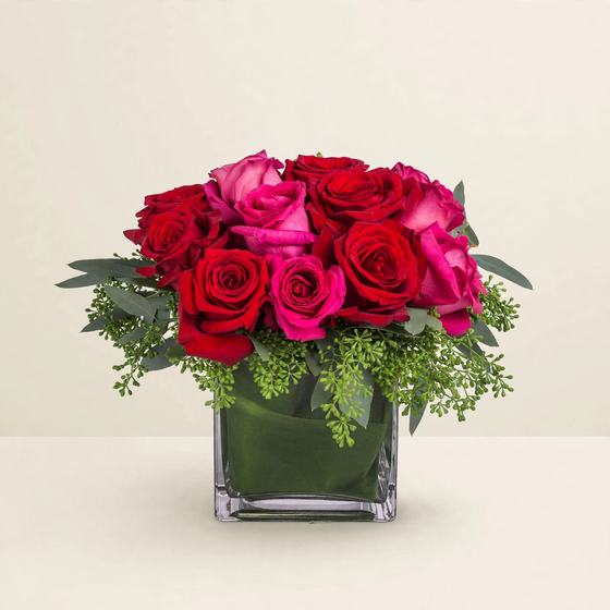 Red and pink roses with fresh greenery arranged in a green cube vase on a cream background