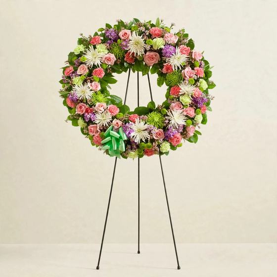 Colorful funeral wreath with pink roses, white spider mums, purple blooms and a green bow on a cream background
