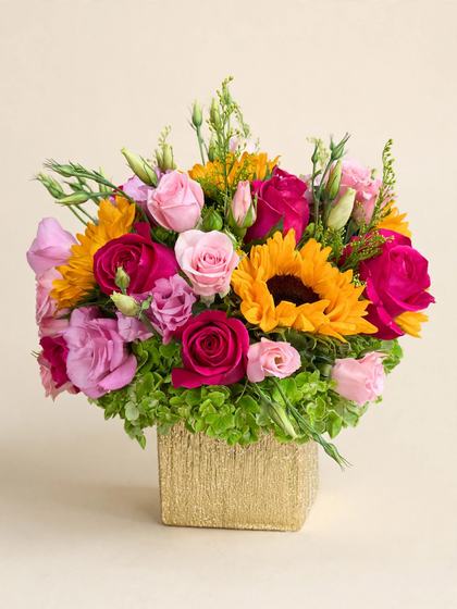Pink and fuchsia roses, sunflowers, lisianthus buds and green hydrangea in a textured gold cube