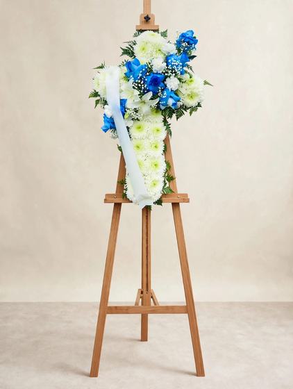 Standing cross spray with blue roses, white lilies, white chrysanthemums and baby's breath on a cream background