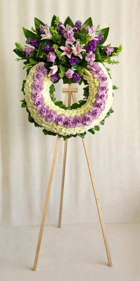 Lavender and pink funeral wreath with lavender roses, pink lilies, purple blooms and white mums on a cream background