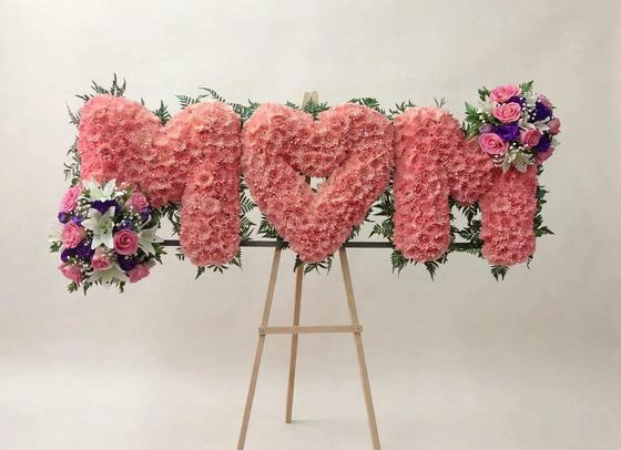 MOM Funeral Letters made of pink cushion daisies with a heart in the center, floral arrangements at both ends, and displayed on a wooden easel