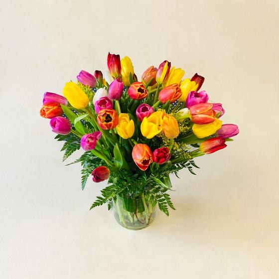 Thirty yellow, pink, orange, red and white tulips arranged with greenery in a clear glass vase