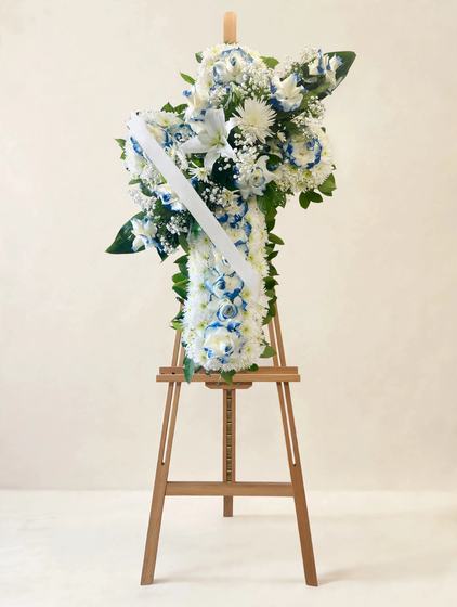 Standing cross spray with white lilies, blue-tipped white roses, white chrysanthemums and baby's breath on a cream background