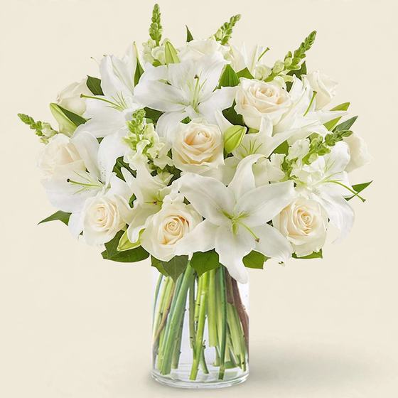 Serene White Bouquet with white lilies and white roses in a clear glass vase on a cream background