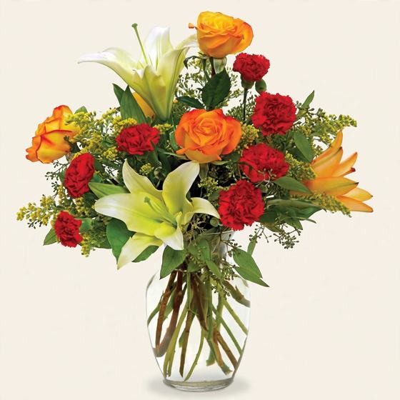 Orange and yellow roses, white lilies and red carnations in a glass vase on a cream background