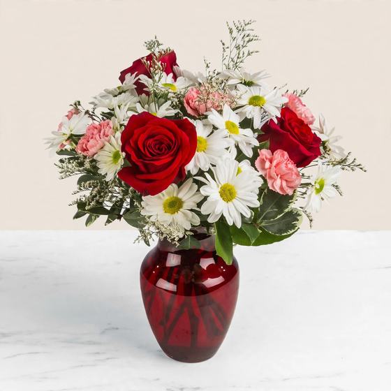 Red roses, pink carnations and white daisies arranged in a red glass vase on a cream background