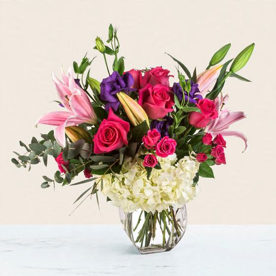 Hot pink roses, pink stargazer lilies, purple lisianthus, white hydrangea, pink spray roses and eucalyptus in a glass vase