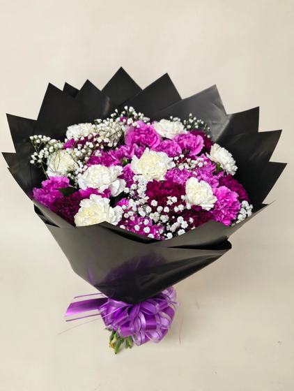 Purple and white carnations with baby's breath in black wrap and a purple bow on a cream background