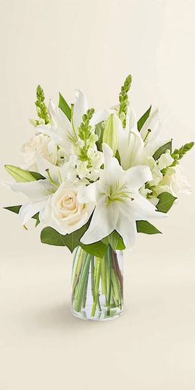White Lily and Rose Tribute with white lilies, white roses and white snapdragons in a clear glass vase on a cream background